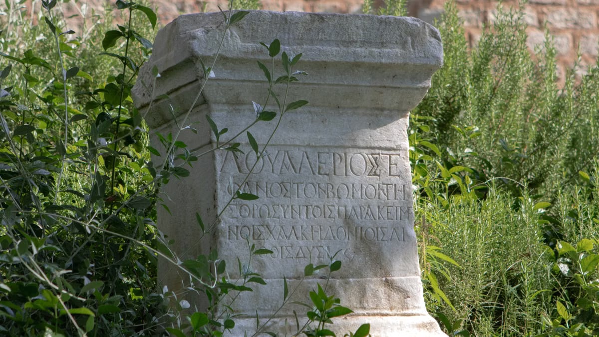Ancient stone monument with Greek inscriptions surrounded by lush greenery, revealing historical ruins.
